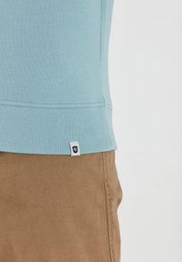 Lichtblauwe katoenen trui met een geribbelde zoom en een klein merketiket aan de zijkant, gecombineerd met beige broek die op de achtergrond zichtbaar is.