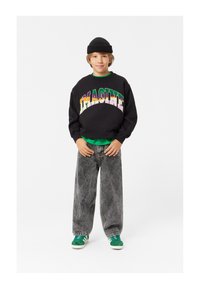 Felpa nera con testo "IMAGINE" multicolore, t-shirt verde sotto, jeans larghi grigi e sneakers verdi con dettagli bianchi.