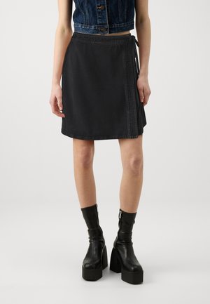 Femme portant une jupe portefeuille noire longueur genou, des bottines noires à plateforme et un gilet court en denim foncé.
