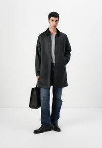 Man wearing a black buttoned coat, gray shirt, blue jeans, black shoes, holding a black leather tote bag, standing against a white background.