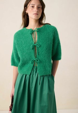 Groene pluizige gebreide cardigan met korte mouwen, voorzien van een keyhole sluiting aan de voorkant, vastgemaakt met drie strikjes. Draagt over een bijpassende groene jurk.