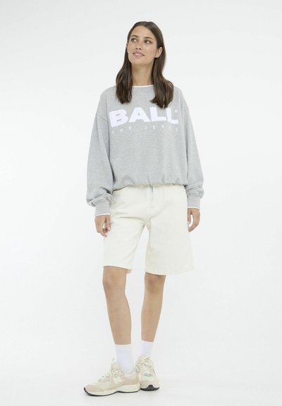 Felpa grigia con testo bianco "BALL" e "THE JEANS", abbinata a pantaloni corti crema e sneakers beige. Il modello si trova davanti a uno sfondo semplice.