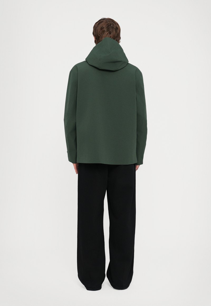 Veste à capuche verte en tissu lisse, présentant une coupe ample et des épaules tombantes, associée à un pantalon noir fluide.