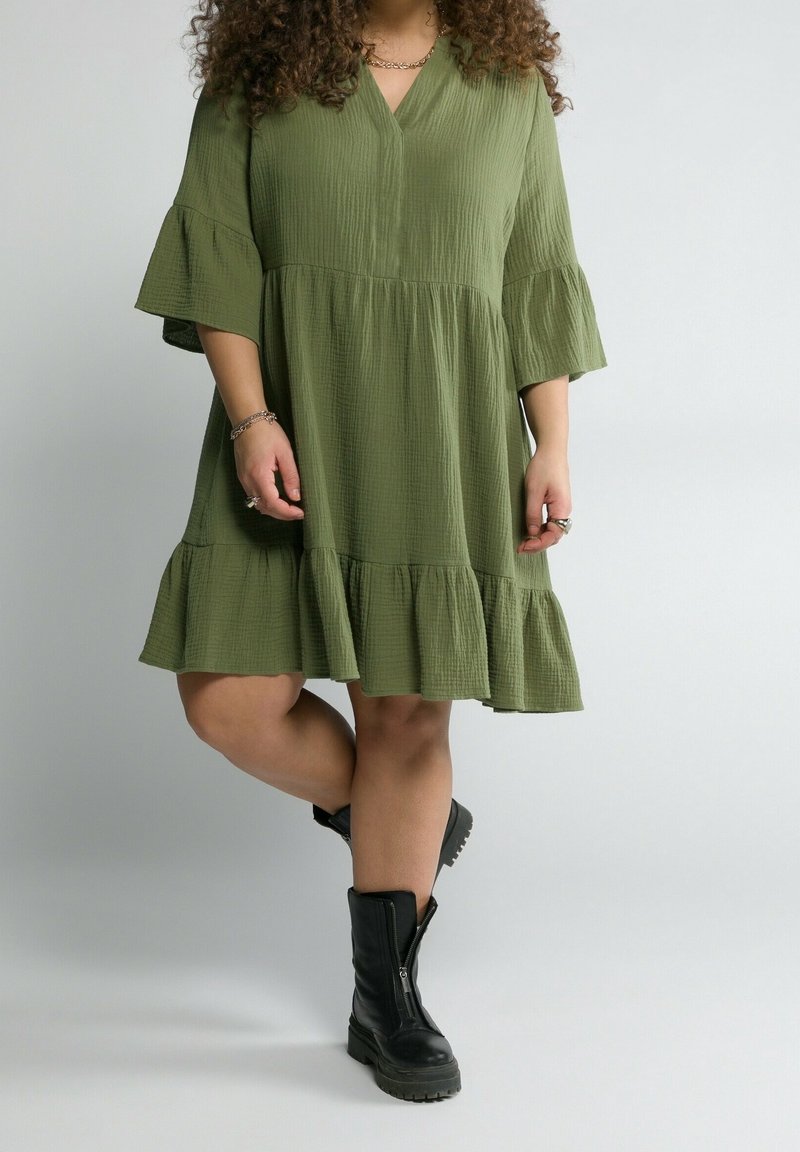 Vestido color verde oliva hasta la rodilla, con cuello en V, mangas acampanadas y dobladillo con volantes en capas; combinado con botines negros con cremallera en el tobillo.