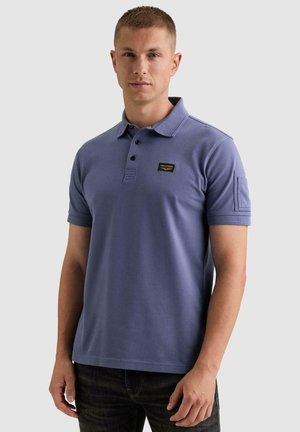 Jeune homme portant un polo bleu à manches courtes avec col boutonné et petit détail de poche sur la manche gauche.