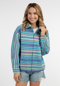 Giacca colorata con un motivo geometrico in turchese, rosa e verde, con zip frontale e vestibilità relax. Abbinata a pantaloni corti di jeans.