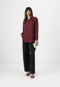 Camicia bordeaux con maniche lunghe, abbinata a pantaloni neri a gamba larga e sandali con tacco neri. Tiene in mano una pochette argentata sfumata.