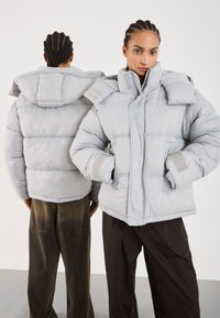 Svijetlo siva puffer jakna s visokim ovratnikom, horizontalnim šavovima i čičak kopčama. Ima velike džepove i opušteni kroj.