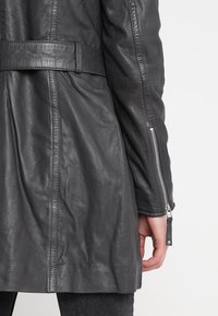 Veste en cuir noir au design structuré, taille ceinturée et poignets zippés, mettant en valeur une texture lisse et une silhouette ajustée.