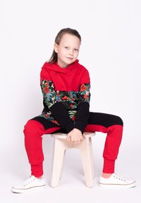 Czerwona i czarna bluza z kapturem w kwiatowy wzór i długimi rękawami, zestawiona z czarno-czerwonymi spodniami. Białe trampki z czerwonymi akcentami. Siedzi na stołku.