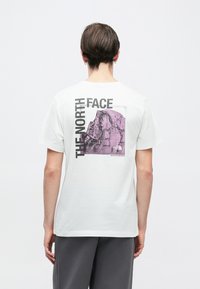 Λευκό βαμβακερό μπλουζάκι με γραφικό μοβ βουνό και το κείμενο "THE NORTH FACE" στην πλάτη. Σχεδιασμός με κοντά μανίκια και στρογγυλή λαιμόκοψη.