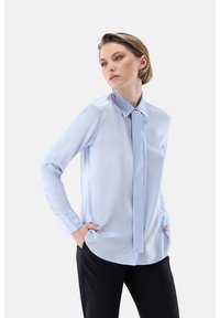 Light blue, long-sleeve blouse in smooth fabric with a collar and a matching tie. Features a rounded hem and loose fit, paired with black slacks.