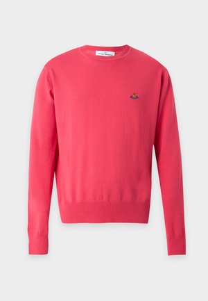 Uomo che indossa una camicia rosa brillante a maniche lunghe, pantaloni neri larghi e scarpe bianche, in piedi davanti a uno sfondo chiaro e uniforme.
