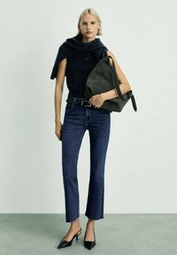 Črna brezrokavnika, modri zvonasti jeans in črne čevlje s špicami. Manekenka drži sivo torbico. Gladka tekstura, minimalističen dizajn.