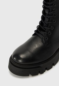Botín de cuero negro con cordones, con punta redondeada, suela gruesa y textura de cuero. Ojetes y cordones visibles en la parte superior.