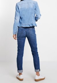 Ljusblå denimjacka med volangnedrekanter och pärlprydnader på ärmarna, matchad med mörkblå jeans och vita sneakers.