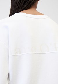 Felpa bianca con collo rotondo, caratterizzata da una texture morbida. Leggera scritta del marchio in colore chiaro sulla parte superiore della schiena.