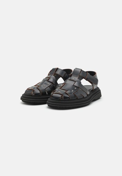 Sandales en synthétique noir avec des brides tissées, des côtés ouverts et des coutures brunes contrastantes. Semelle épaisse noire avec une surface texturée.