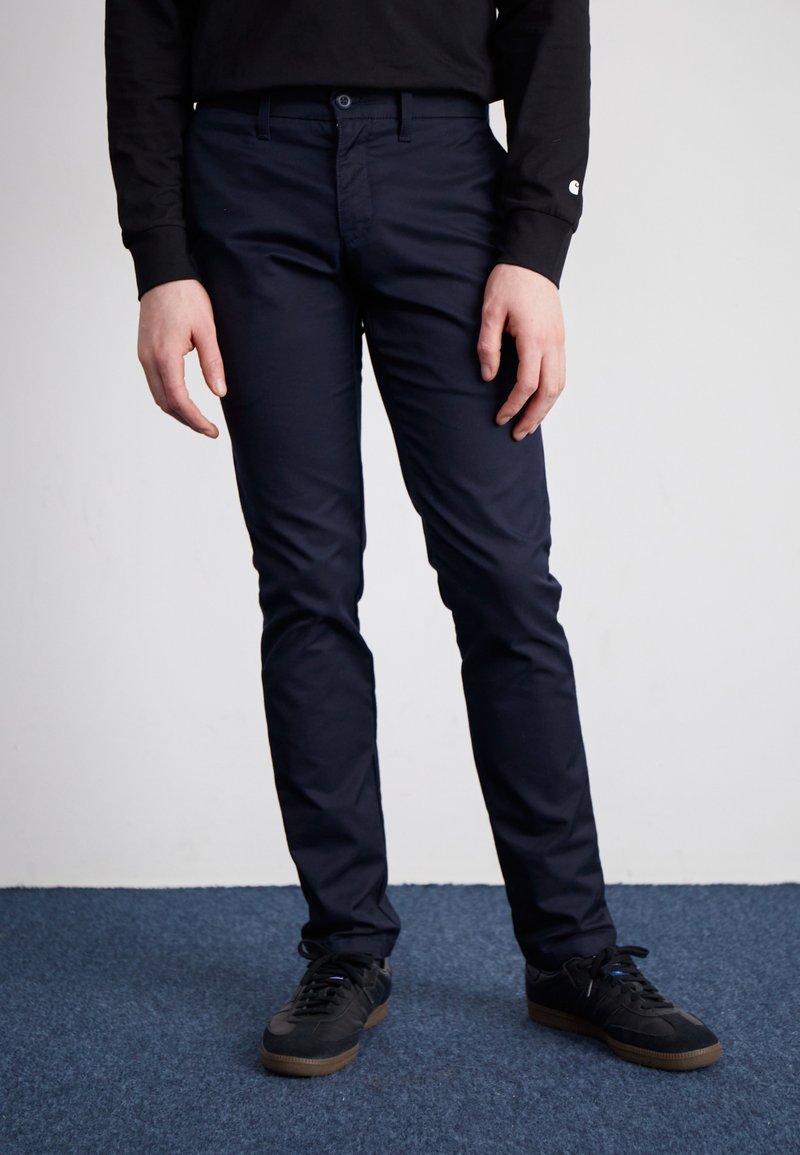 Pantalon slim-fit bleu marine avec une taille plate, fermeture à bouton et poches ; porté avec des baskets noires sur une surface de moquette bleue.