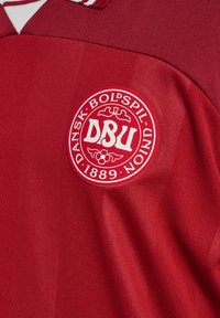 Rotes Sporttrikot mit einem auffälligen, runden, gestickten Patch, auf dem "DBU" und "DANSK BOLSPIL UNION" in weißem Text steht, aus glattem Stoff gefertigt.