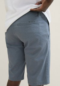 Hellblaue, strukturierte Shorts mit geradem Schnitt, die eine Gesäßtasche und ein Logodetail aufweisen. Der Stoff wirkt atmungsaktiv und leicht.