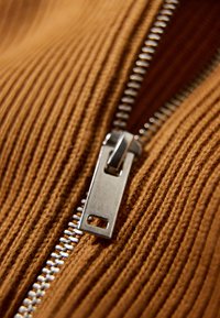 Tessuto rigato marrone con una zip metallica. La zip presenta un cursore e dei denti argentati, che completano la texture del materiale.