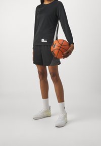 Camiseta deportiva de manga larga negra con detalles blancos, combinada con pantalones cortos de malla negra. Sosteniendo un balón de baloncesto naranja, usando zapatillas blancas y calcetines.