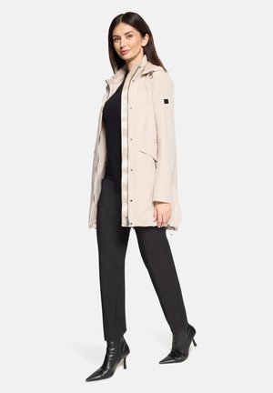 Beige waterproof jacket with a zipper front, adjustable hood, and side pockets, paired with black fitted pants and black pointed boots.