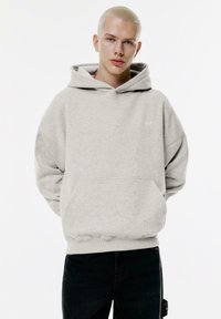 Hoodie oversize grigio chiaro realizzato in tessuto morbido, dotato di tasca frontale e cappuccio con coulisse. Logo minimalista sul petto.