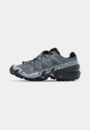 Grauer Trailrunning-Schuh mit strukturiertem Obermaterial, schwarzen Akzenten, niedriger Silhouette und multidirektionalen Stollen auf der Sohle für besseren Halt.