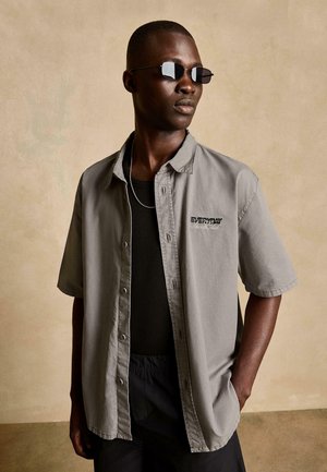 Muž, ktorý má na sebe čierne slnečné okuliare, sivú krátkoročnú košeľu na gombíky ponad čierny top, strieborný náhrdelník, stojí s jednou rukou v vrecku.