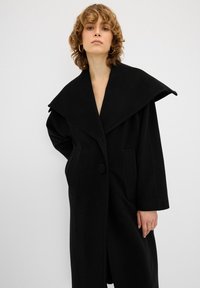 Cappotto di lana nera con un grande e ampio colletto, maniche lunghe e due tasche anteriori. Presenta una chiusura con un solo bottone e una silhouette rilassata.
