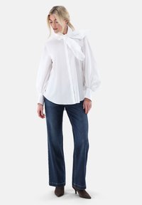 White blouse with a large bow tie at the collar, puffed sleeves, and a relaxed fit, paired with dark blue wide-leg jeans and brown heels.
