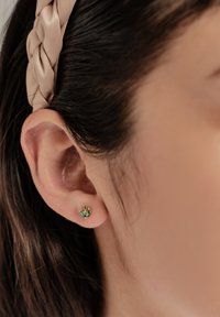 Pendientes de stud en tono dorado con un diseño floral que presenta dos cristales verdes. Forma sencilla con un acabado pulido. Llevados en la oreja derecha.