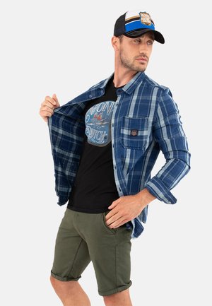 Homme portant une chemise à carreaux bleue par-dessus un t-shirt graphique noir, un short vert olive et une casquette trucker noire et bleue, posant avec la chemise ouverte.