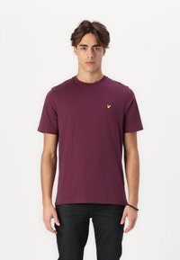 T-shirt à manches courtes de couleur bordeaux en coton doux, avec un col rond et un petit logo jaune sur la poitrine gauch.