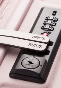 La cerniera e il lucchetto combinato della valigia Bric's in argento, posizionati su uno sfondo testurizzato di rosa chiaro, caratterizzati da un meccanismo di chiusura approvato dalla TSA.