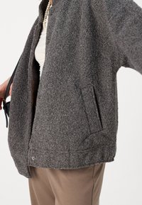 Grå, textureradulljacka med tryckknappar, sidofickor och en avslappnad passform, parat med ljusa byxor.