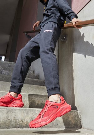 Personne portant un pantalon de survêtement foncé et des baskets HUGO rouge vif descendant des escaliers en béton, tenant une rambarde en métal.