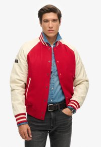 Red and cream varsity jacket with ribbed cuffs and collar, featuring a snap closure and side pockets. Worn over a denim shirt.