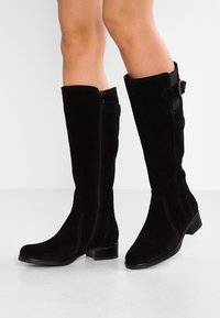 Bottes en daim noir montant jusqu'au genou, avec un talon plat et une fermeture éclair latérale. Présentent un design simple avec une texture lisse et des détails de couture distinctifs.