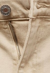 Beige denimstof met zichtbare stiksels, een zilverkleurige metalen rits en een drukknoopsluiting aan de voorkant van de tailleband.