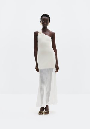 Femeie purtând o rochie maxi crem cu un umăr gol, cu cataramă aurie și fustă inferioară plisată și transparentă, asortată cu sandale și cercei aurii.