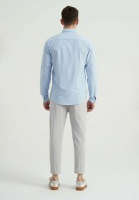 Chemise à manches longues bleu clair avec un ourlet arrondi, associée à un pantalon gris clair ajusté et des chaussures beiges avec des accents blancs.