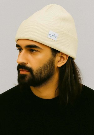 Cream ribbed beanie made of knitted fabric, featuring a small white label with a blue logo accent, worn with a black sweater.