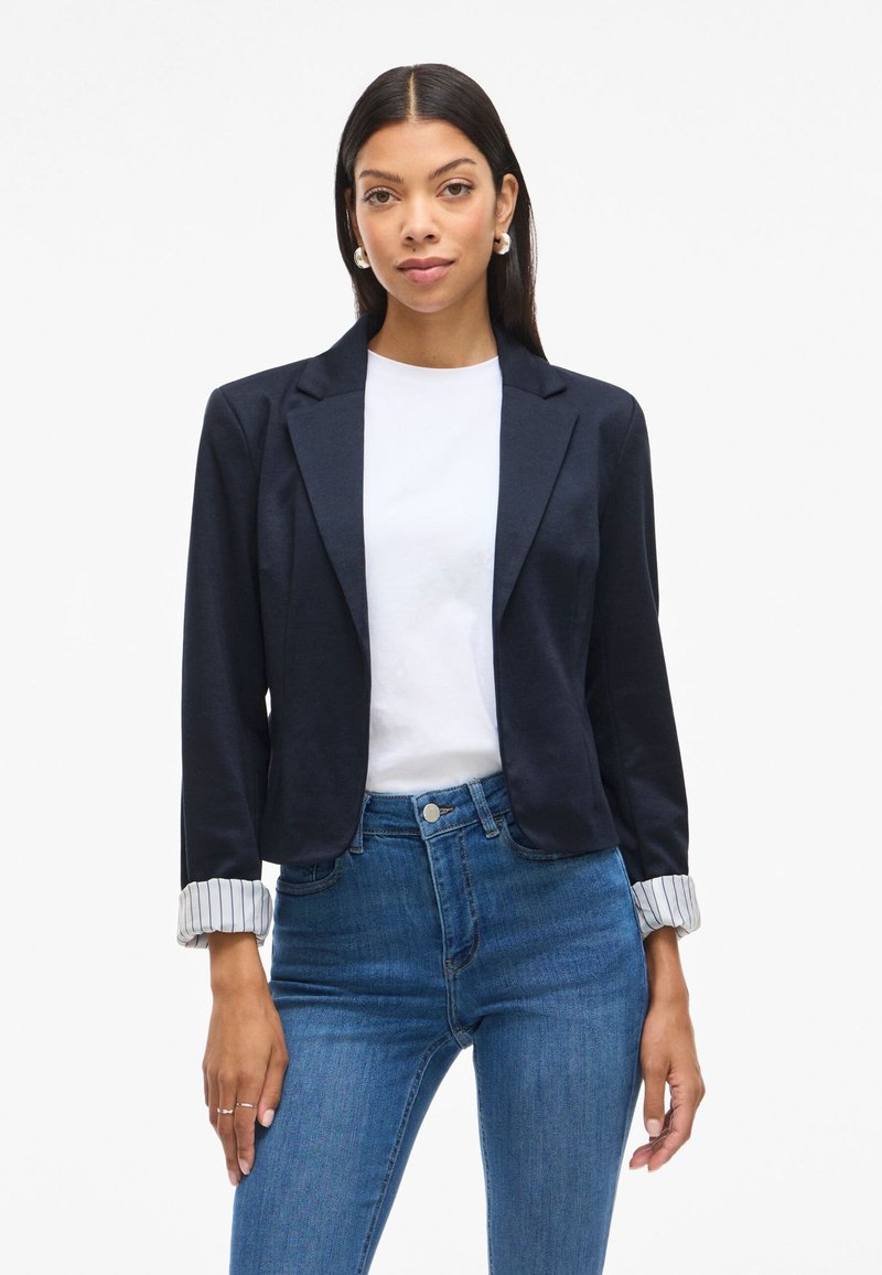 Tamsiai mėlynas švarkas, pritaikytas prie figūros, su atlenkamu apykakle ir dryžuotais rankogaliais. Dėvimas virš balto marškinėlio ir mėlynų džinsų.