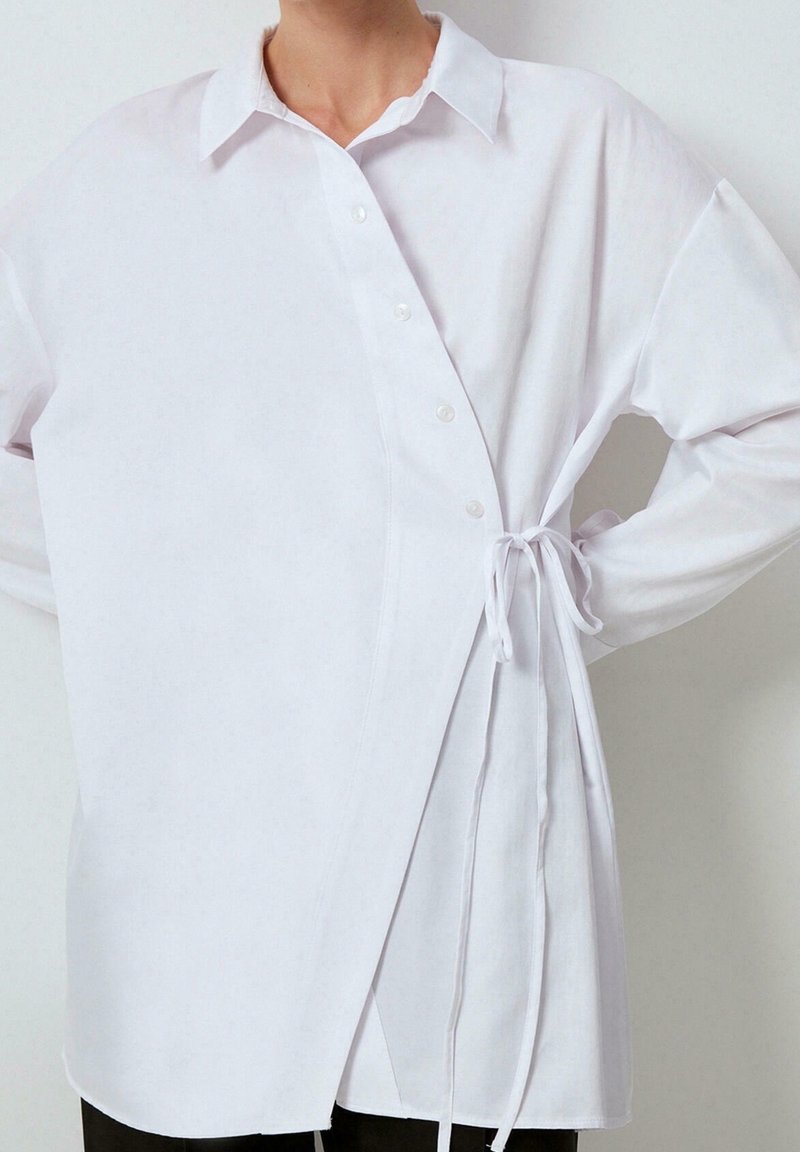 Personne portant une chemise croisée blanche à manches longues avec liens sur les côtés et boutons, debout les mains sur les hanches devant un fond uni.