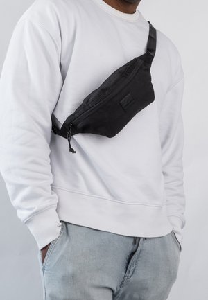 Person wearing a white sweatshirt and light jeans with a black crossbody waist bag fastened across the chest.