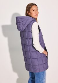 Lila gesteppte Weste mit Kapuze, ausgestattet mit Seitentaschen, einem strukturierten Finish und einer lässigen Passform, getragen über einem weißen Langarmhemd.