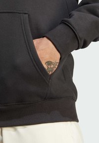 Felpa nera con tasca a marsupio, polsini e orlo a costine, che presenta un tessuto testurizzato. Mano parzialmente visibile con un tatuaggio.